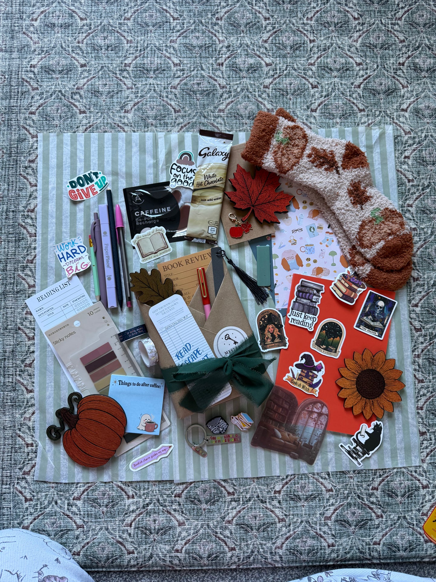 A styled flat-lay of a fall-themed gift set on a striped fabric mat, featuring a brown and cream knit stocking, autumn stickers, a pumpkin patch, a sunflower cutout, a green bow, and various stationery items on a textured gray background.
