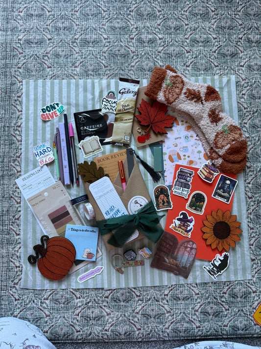 A styled flat-lay of a fall-themed gift set on a striped fabric mat, featuring a brown and cream knit stocking, autumn stickers, a pumpkin patch, a sunflower cutout, a green bow, and various stationery items on a textured gray background.